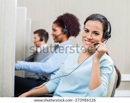Portrait of smiling female customer service representative wearing headset with colleagues working in background at office