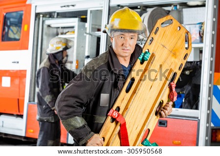 Confident male firefighter holding wooden stretcher against truck at fire station