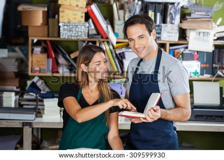 Portrait of happy male worker holding book while colleague pointing at it in factory