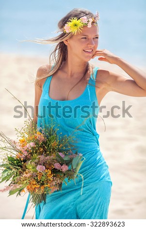 One young woman with blond hair in long blue dress, with bunch of flowers in hands and flower wreath on head, standing on beach in sunny day.