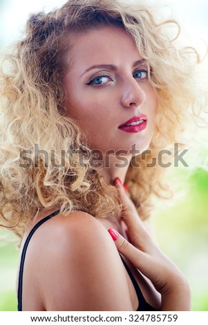 Portrait of young woman with blond curly hair, red lips and black eyeliner, hands with red nails, outdoors on sunny day