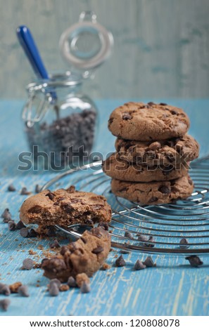 Side closeup view of cookies with chocolate drops on round iron stand, jars with chocolate drops on cracked blue background