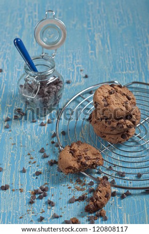 Top closeup view of broken cookie and biscuits with chocolate drops on round iron stand, jars with chocolate drops on cracked blue background