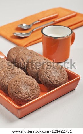 Closeup side view of biscuits in orange ceramic pan, spoons on napkin and cup of milk on white wooden background