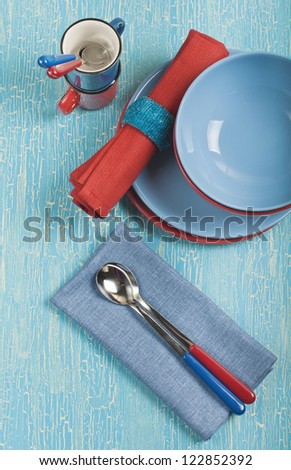 Top view of the kitchen utensils: red and blue plates, cups, spoons and napkins on a blue cracked background