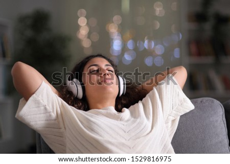 Relaxed tenant wearing headphones resting listening to music sitting on a couch in the living room in the night at home