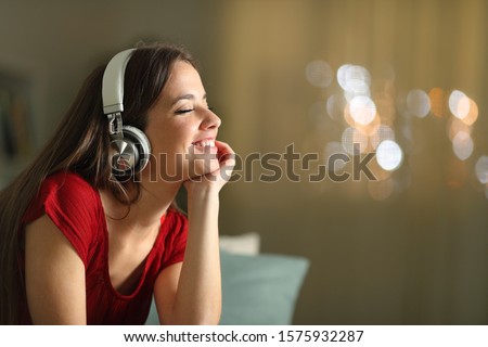 Happy woman wearing wireless headphones listening to music in the night sitting on a couch in the living room at home