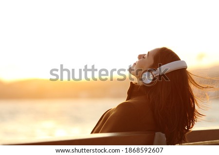 Woman wearing headphones listening to music breathing fresh air relaxing sitting on a bench in winter on the beach