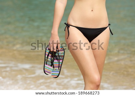 Close up of a woman hips holding flip flops on the beach with the water in the background