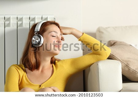 Relaxed woman sitting leaning on radiator listening to music with headphones at home