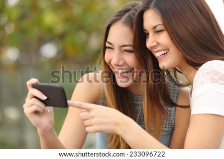 Two funny women friends laughing and sharing social media videos in a smart phone outdoors