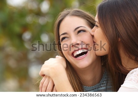 Woman laughing with perfect teeth while a friend is kissing her with a green background