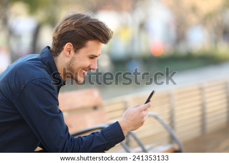 Profile of a happy guy using a smart phone sitting on a bench in a park