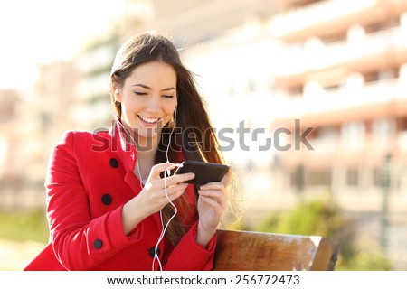 Woman watching videos in a smart phone with earphones sitting in a park with an unfocused buildings in the background
