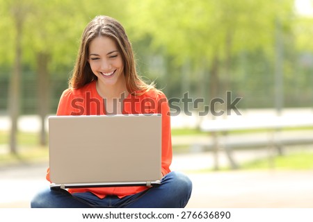 Front view of a happy student girl working with a laptop in a green park of an university campus
