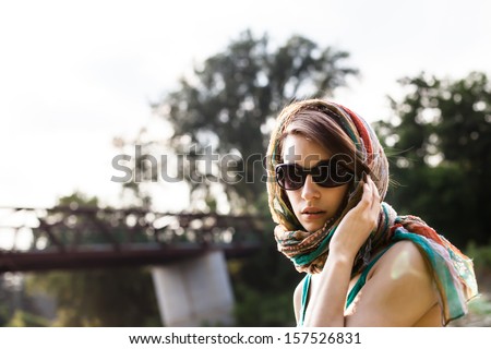 Young woman wearing a teal scarf and sunglasses near a river and natural setting.   She is serene and thoughtful.
