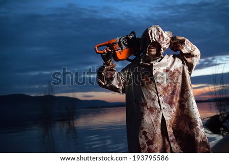 Maniac with the chainsaw dressed in a dirty bloody raincoat. Sunset river on the background