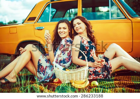 Two beautiful women on the picnic are drinking wine near a car.