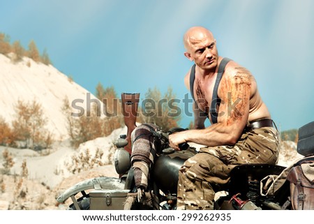 Muscular bald biker with the rifle. Apocalypse background. Instagram-like toning.