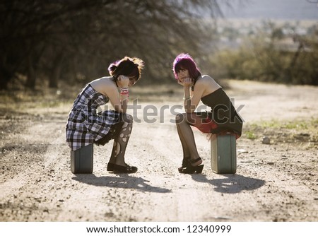Two punk women sitting on suitcases and waiting on a rural road