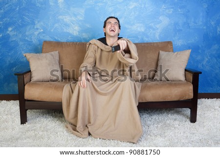 Young Caucasian man in blanket with remote control laughs out loud