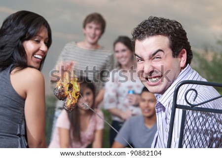 Man with burnt marshmallows on skewer cringes as friends laugh
