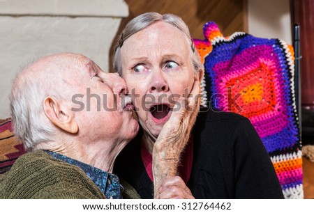 Older gentleman kissing older shocked woman on cheek in livingroom