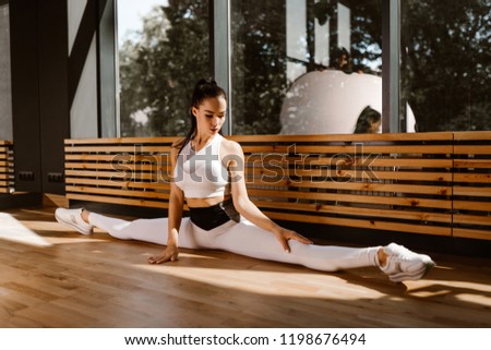 Young slender dark-haired girl dressed in white sports top and tights is doing the Woman doing stretching. Do the splits. Slender young woman doing ...