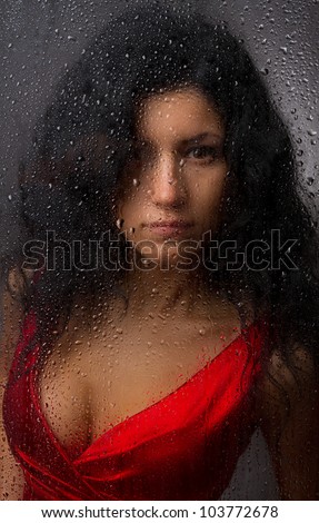 Beautiful girl in a red dress, standing behind a wet glass on a dark background.
