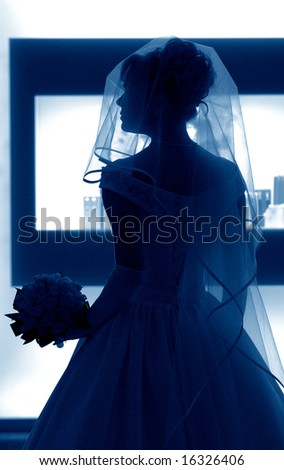 Silhouette of a beautiful bride in a traditional white wedding dress, stood by window with veil and bouquet.