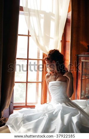 Young adult bride wearing white wedding dress sat by window in sunlight.