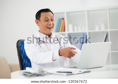 Portrait of amazed doctor sitting in his office and pointing at laptop