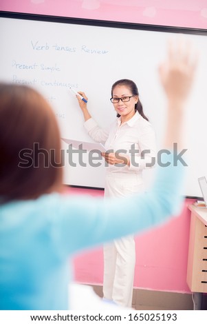 Vertical portrait of a teacher explaining English tenses while one of the pupils put up his hand to ask something on the foreground