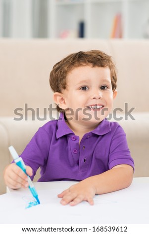 Vertical portrait of a little cheerful boy smiling and looking at camera while painting at home