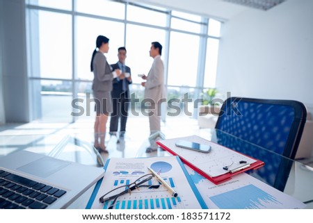 Image of business equipment on the foreground, businesspeople standing and talking with cups of coffee on the background
