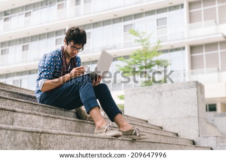 Young man sitting on the stone stairs with laptop and reading a message on the phone