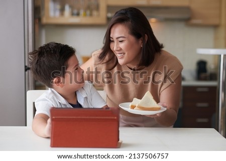Smiling mother bringing plate with sandwiches to little son watching educational video on digital tablet 商業照片 © 