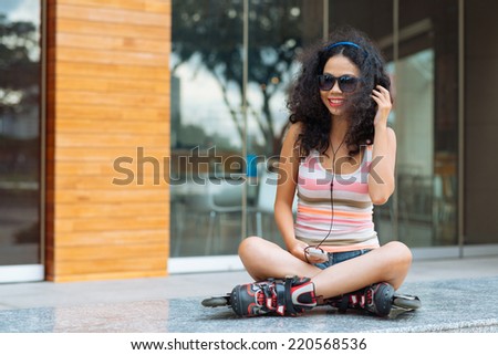 Smiling girl in roller skates sitting outdoors and listening to the music in her phone