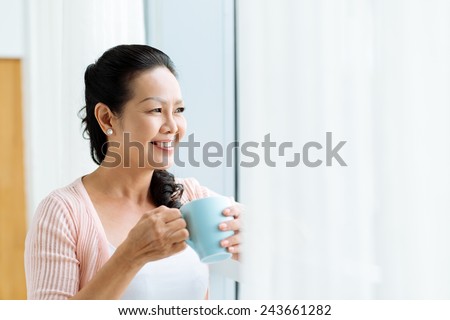 Smiling mature Asian woman standing at the window with a cup of tea