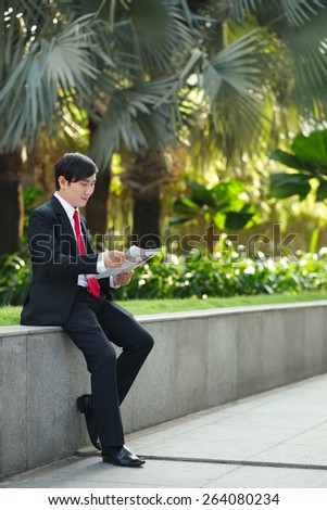 Full-length portrait of Asian businessman reading newspaper outdoors