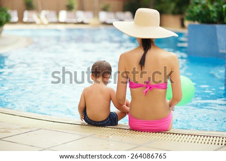 Rear view of mother and son sitting on the edge of the swimming pool: family vacation concept