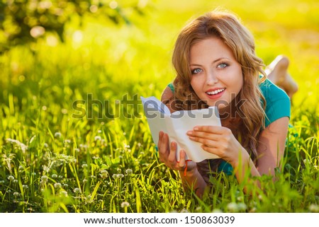 young blond woman reading a book lying on the grass in the shade, she is looking at camera