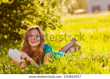 education concept - young blond woman reading a book lying on the grass in the shade