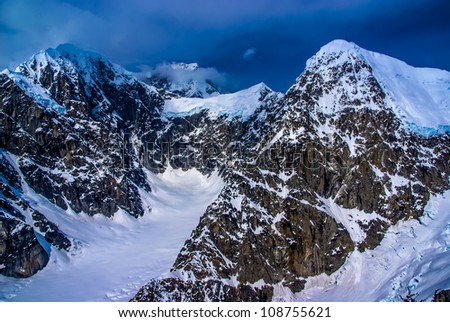 Blue Ice Glacier Headwaters and Snow-pack on Craggy Mountain Peaks with Dark Ominous Clouds.  Aerial View of Denali National Park, Alaska.  A Beautiful Wilderness Snowscape of Rock, Snow, and Ice.
