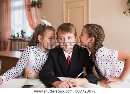 The girls whisper in the ear of the boy. Pupils. Smiling. Sitting at a desk.