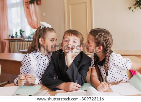 The girls whisper in the ear of the boy. Pupils. Smiling. Sitting at a desk.