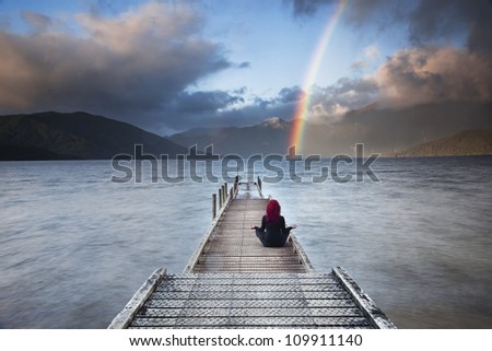 Red haired woman meditating in yoga position on pier overlooking mountains,lake and rainbow