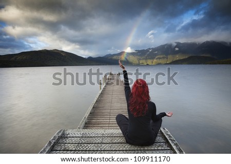 Red haired woman sitting in yoga position on the pier is catching rainbow over the lake