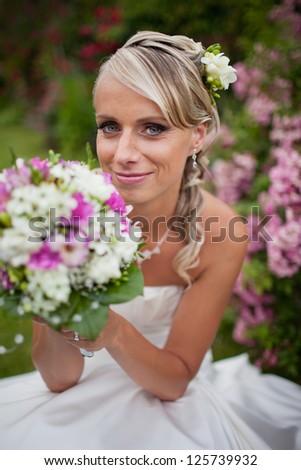 Portrait of blonde smilling happy bride with wedding bouquet of cream roses and pink lilies with pink roses in the background.