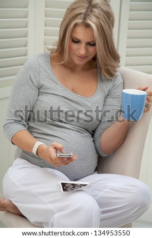 Attractive pregnant woman sitting at home on chair holding blue mug and texting, with ultrasound scan on her lap